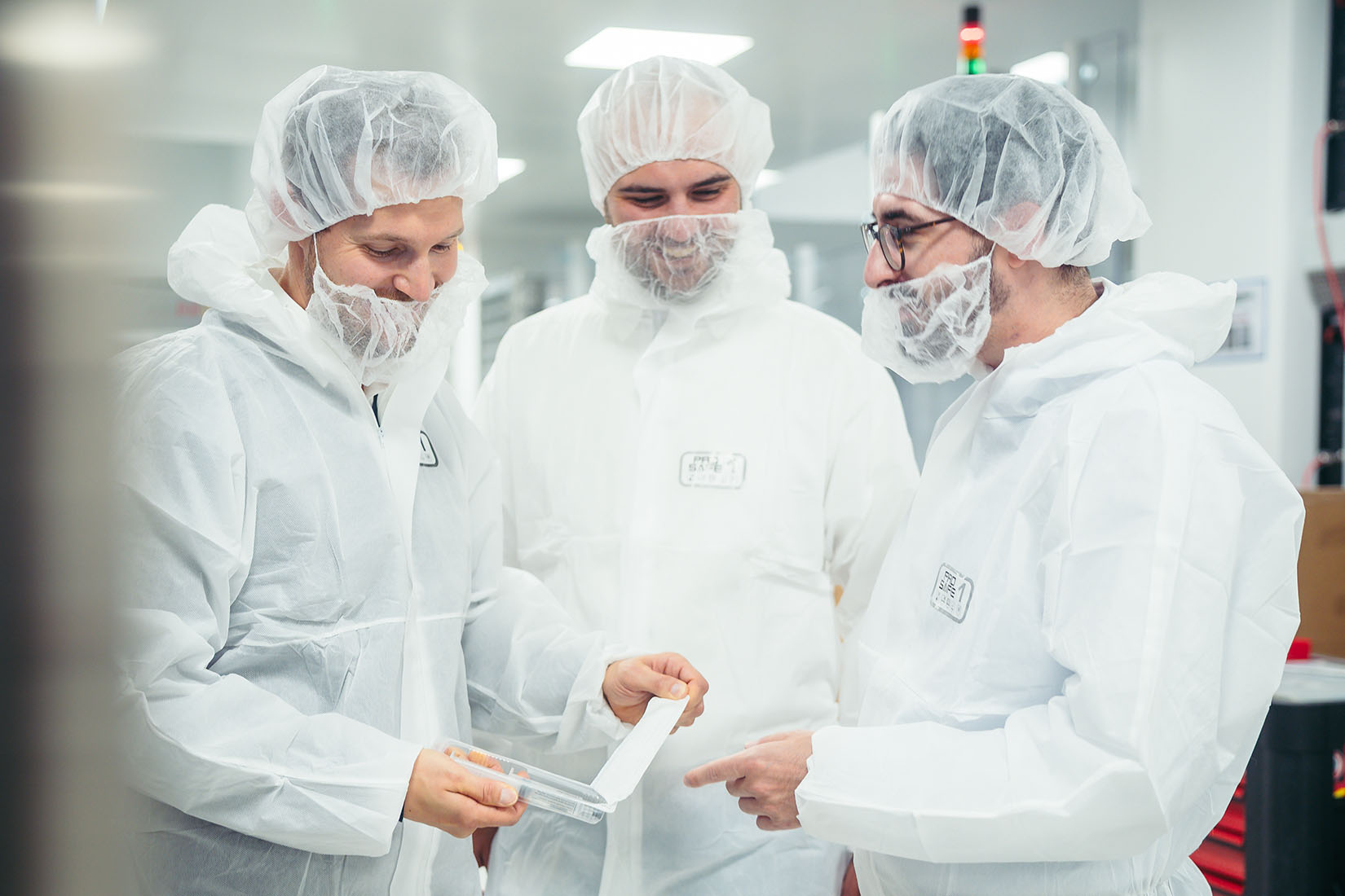 Three men in white lab coats and hairnets discuss a document while examining a transparent container in a cleanroom environment.