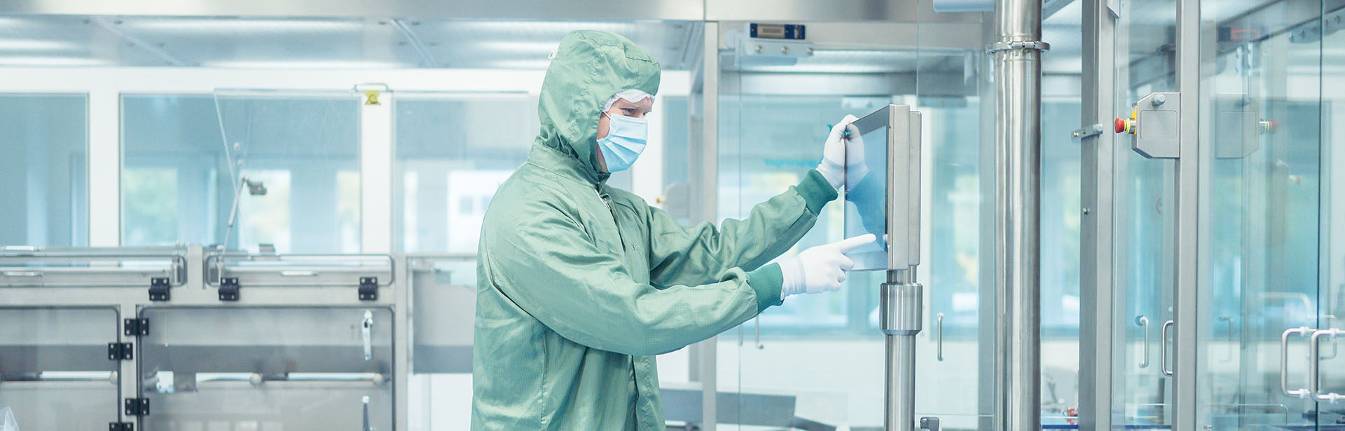 A worker in a green protective suit and mask adjusts equipment in a clean, modern laboratory filled with stainless steel machinery.
