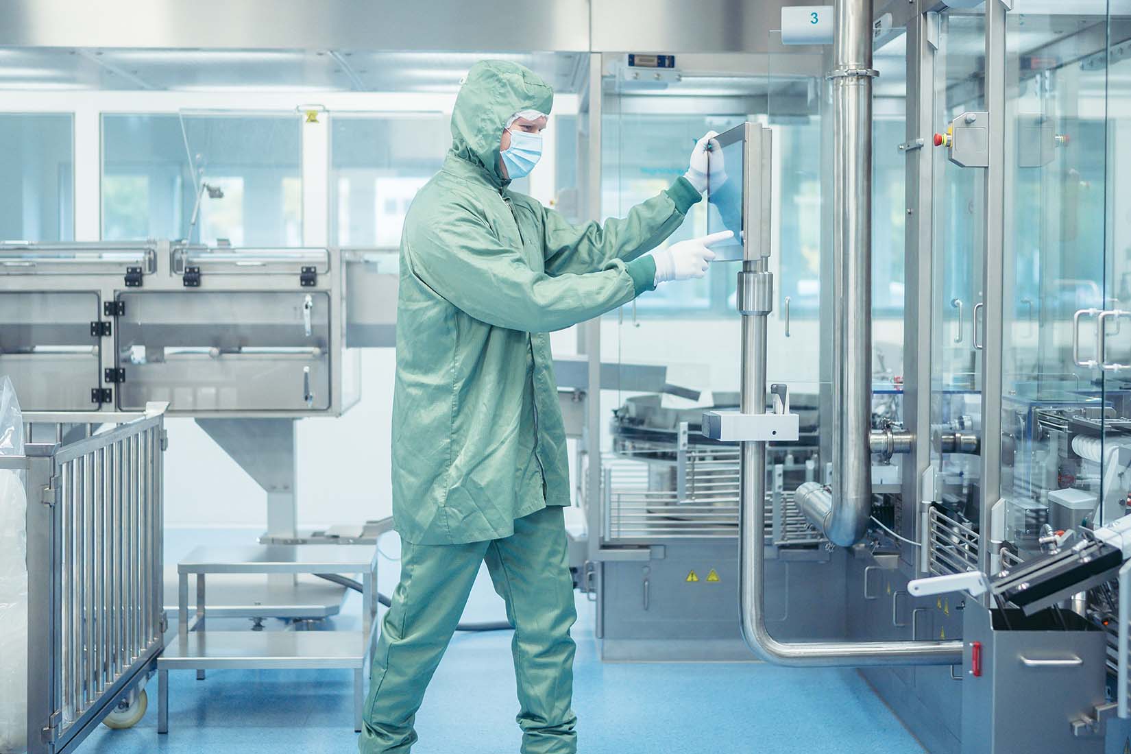 A worker in a green protective suit and mask adjusts equipment in a clean, modern laboratory filled with stainless steel machinery.