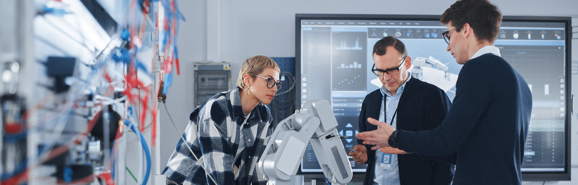 Three professionals engage in a discussion about a robotic arm in a modern tech lab, surrounded by screens displaying data.