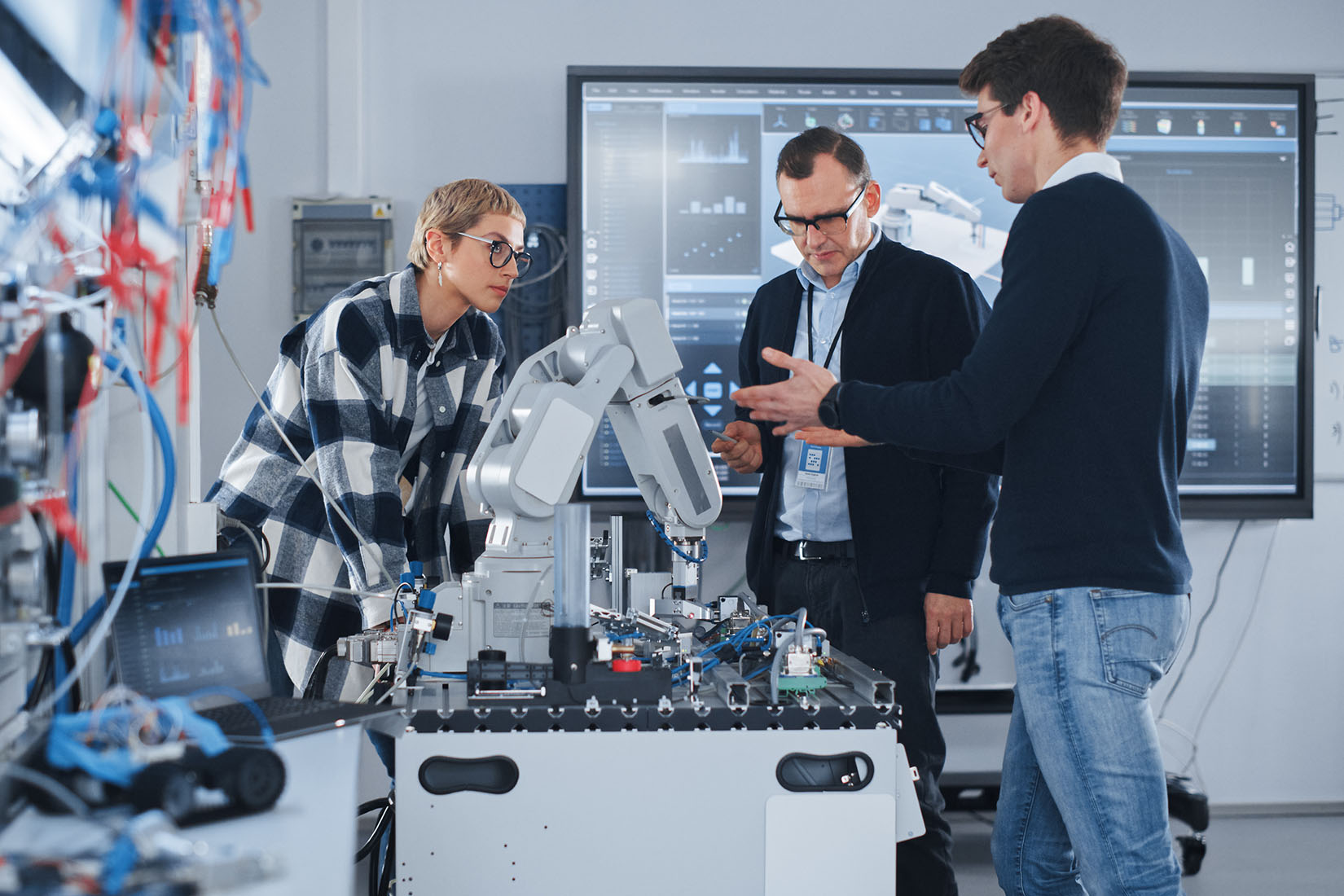 Three professionals engage in a discussion about a robotic arm in a modern tech lab, surrounded by screens displaying data.
