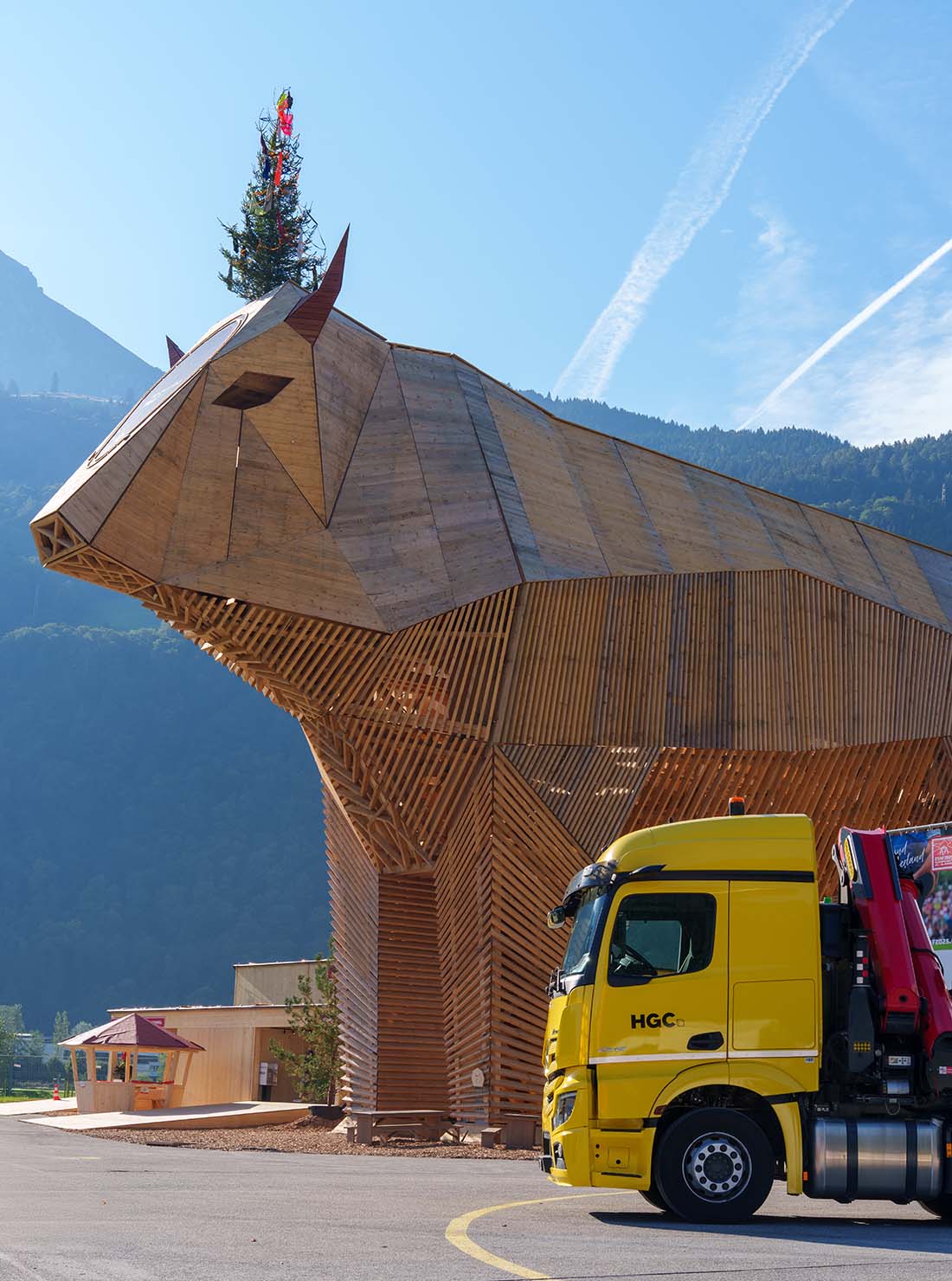 A bright yellow truck is parked near a modern wooden structure, surrounded by lush greenery and mountains in the background.