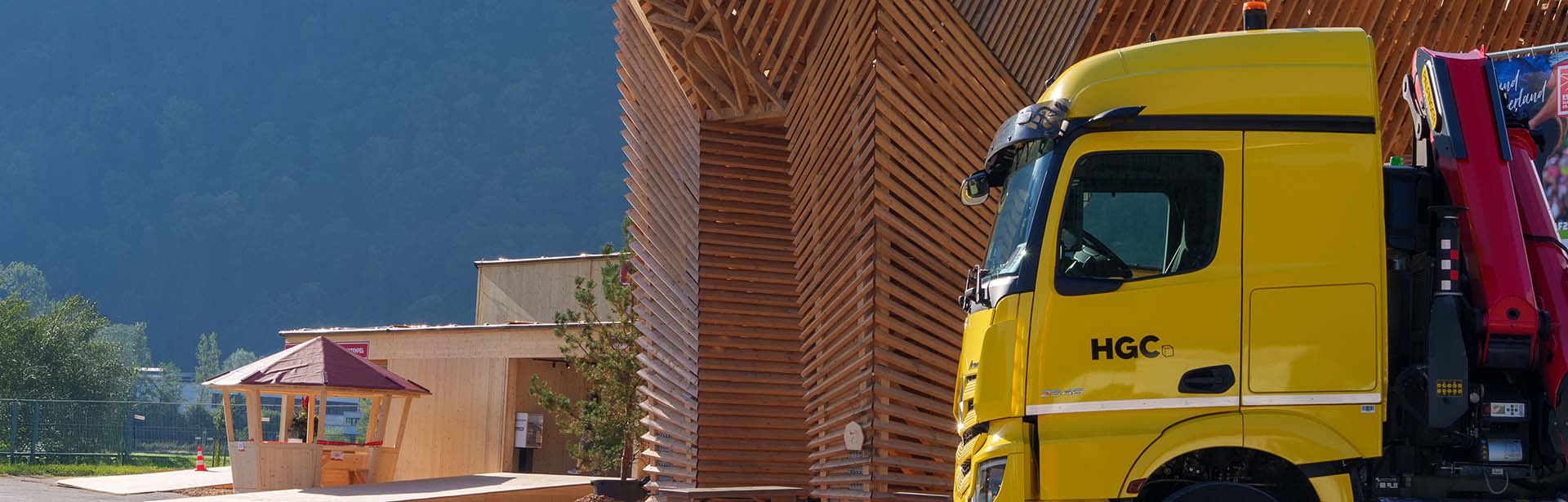 A bright yellow truck is parked near a modern wooden structure, surrounded by lush greenery and mountains in the background.