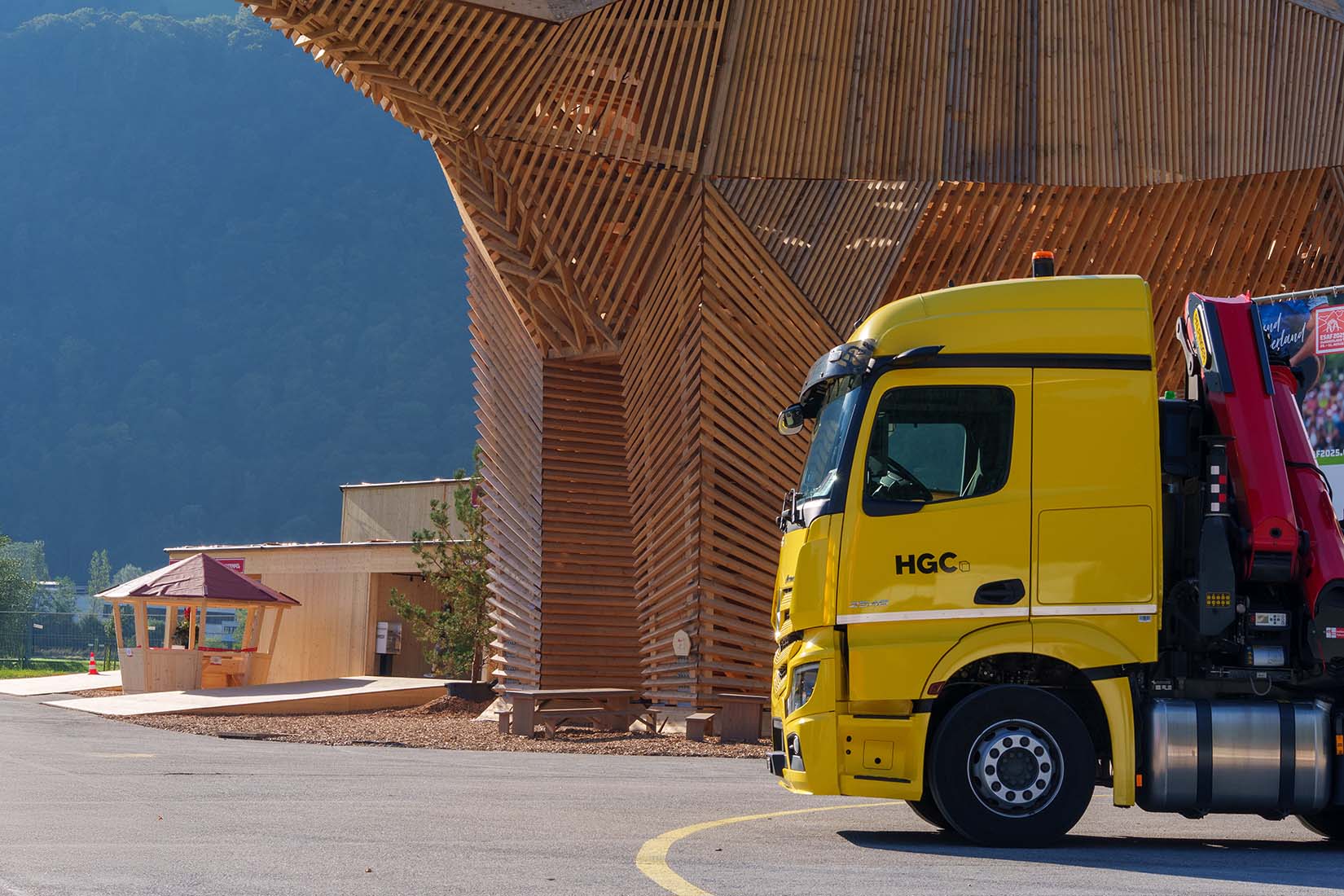 A bright yellow truck is parked near a modern wooden structure, surrounded by lush greenery and mountains in the background.