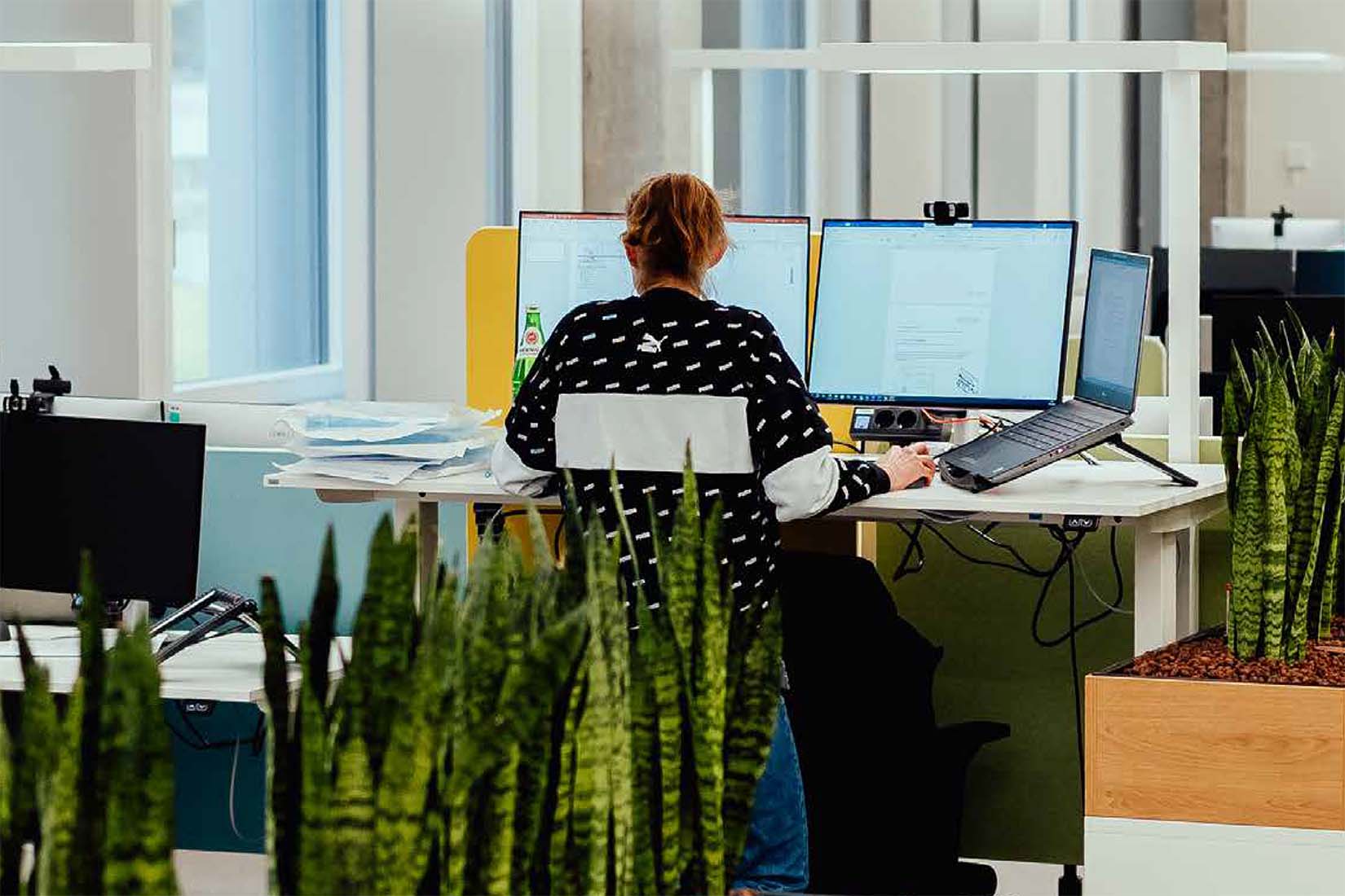 A modern office scene features a woman at her desk, immersed in work, with colorful screens and greenery enhancing the environment.