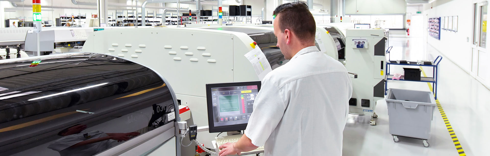 In a bright factory, a worker monitors equipment settings on a computer screen, surrounded by advanced production machines.
