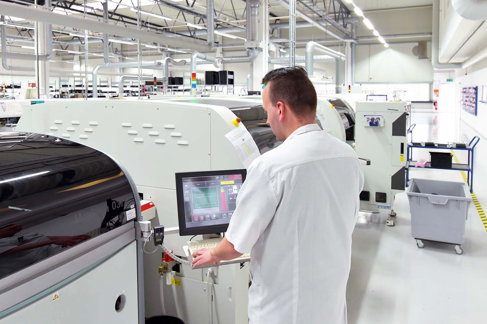 In a bright factory, a worker monitors equipment settings on a computer screen, surrounded by advanced production machines.