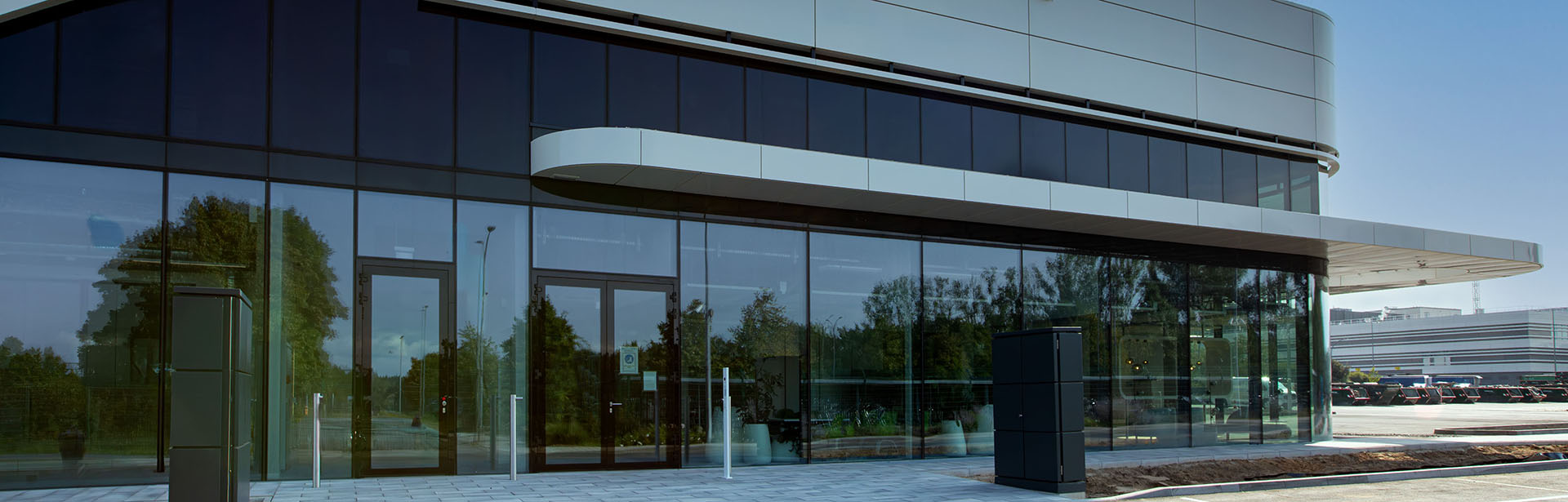 A modern building features sleek glass walls and a curved roof, surrounded by greenery under a clear blue sky.