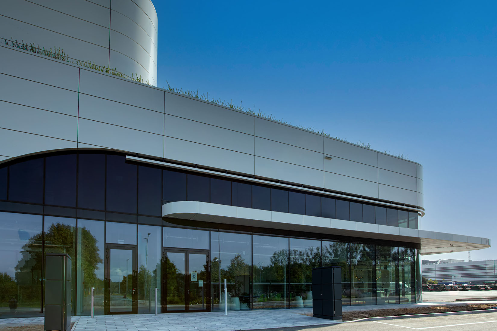 A modern building features sleek glass walls and a curved roof, surrounded by greenery under a clear blue sky.