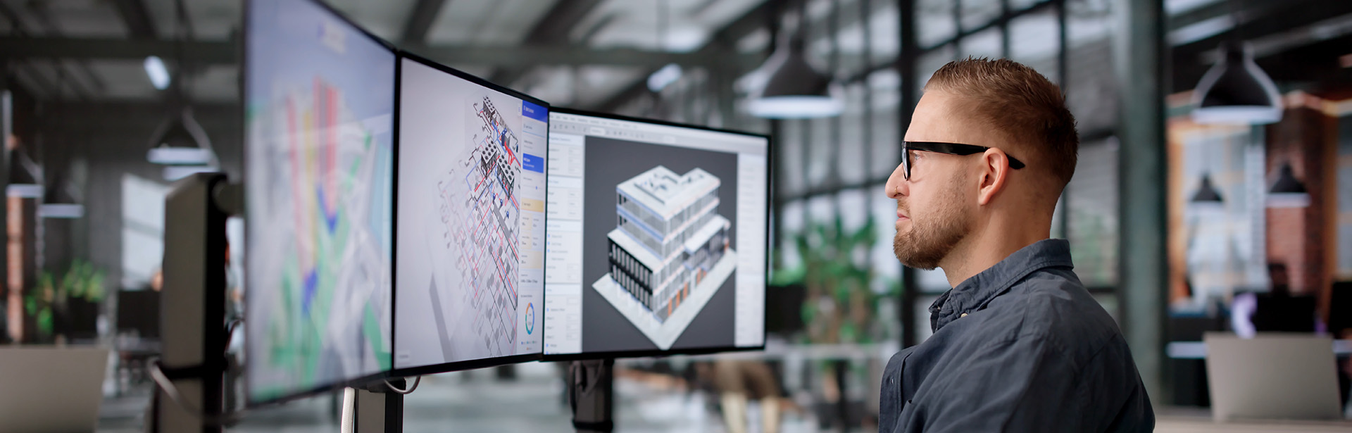 A focused man with glasses analyzes complex architectural designs on three monitors in a modern office environment.