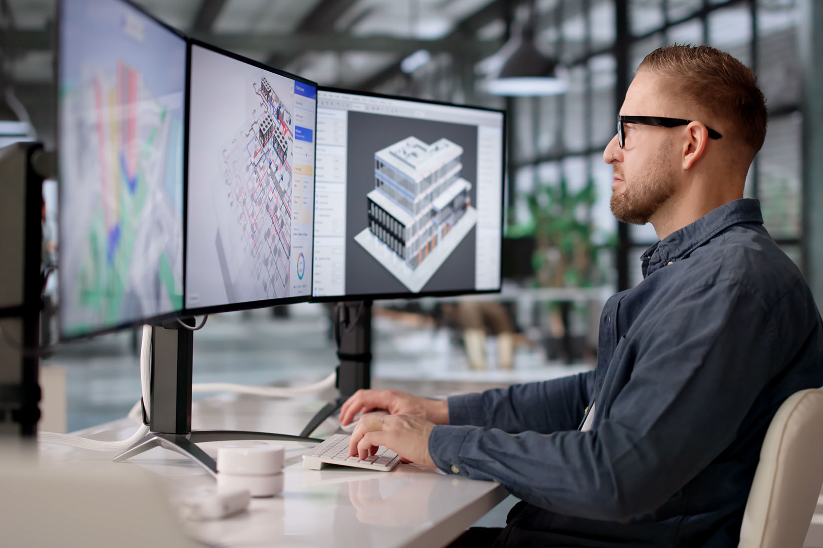 A focused man with glasses analyzes complex architectural designs on three monitors in a modern office environment.