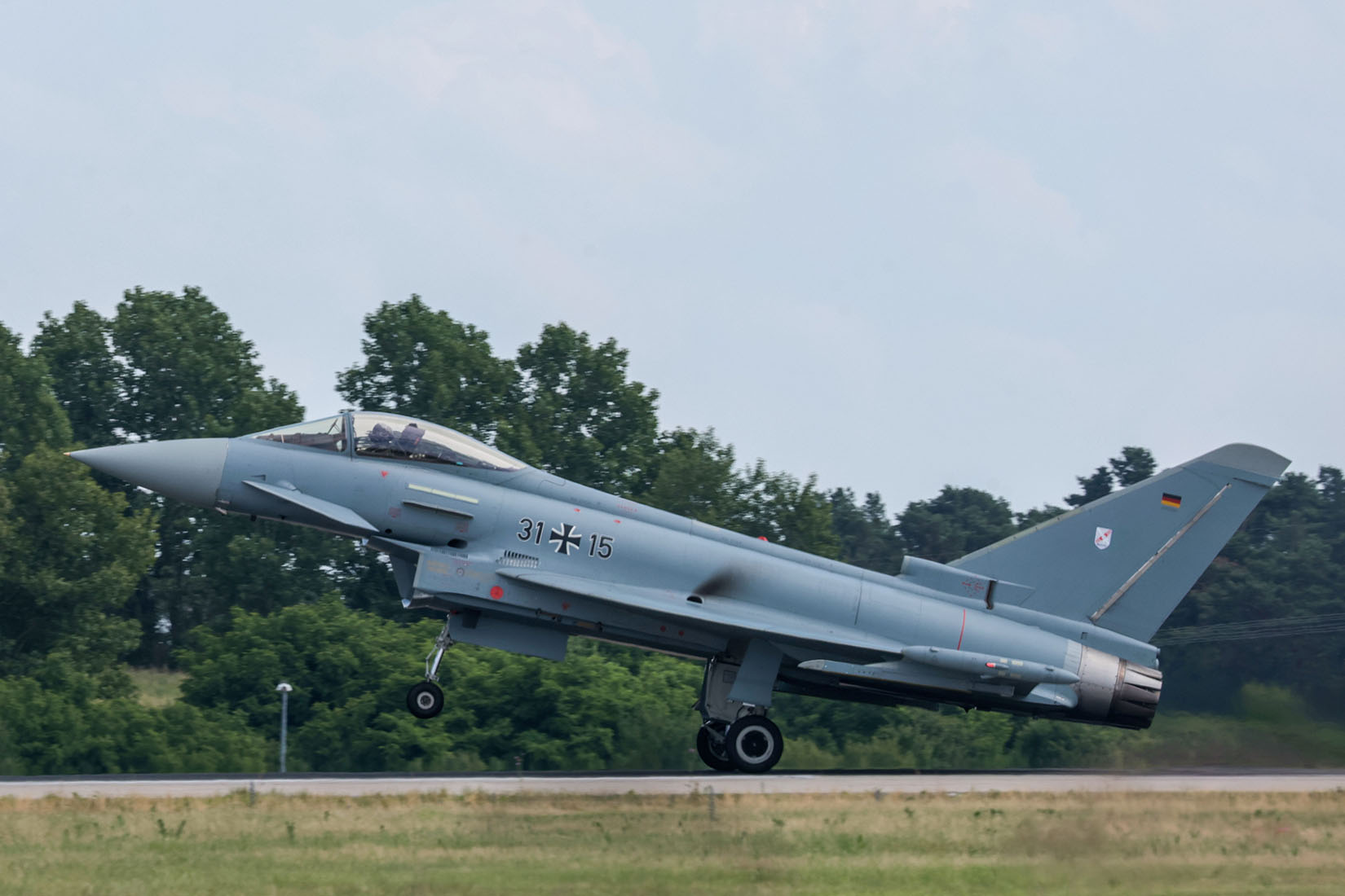 The Eurofighter Typhoon aircraft lifts off the runway, its landing gear retracting as it ascends into the sky, surrounded by nature.