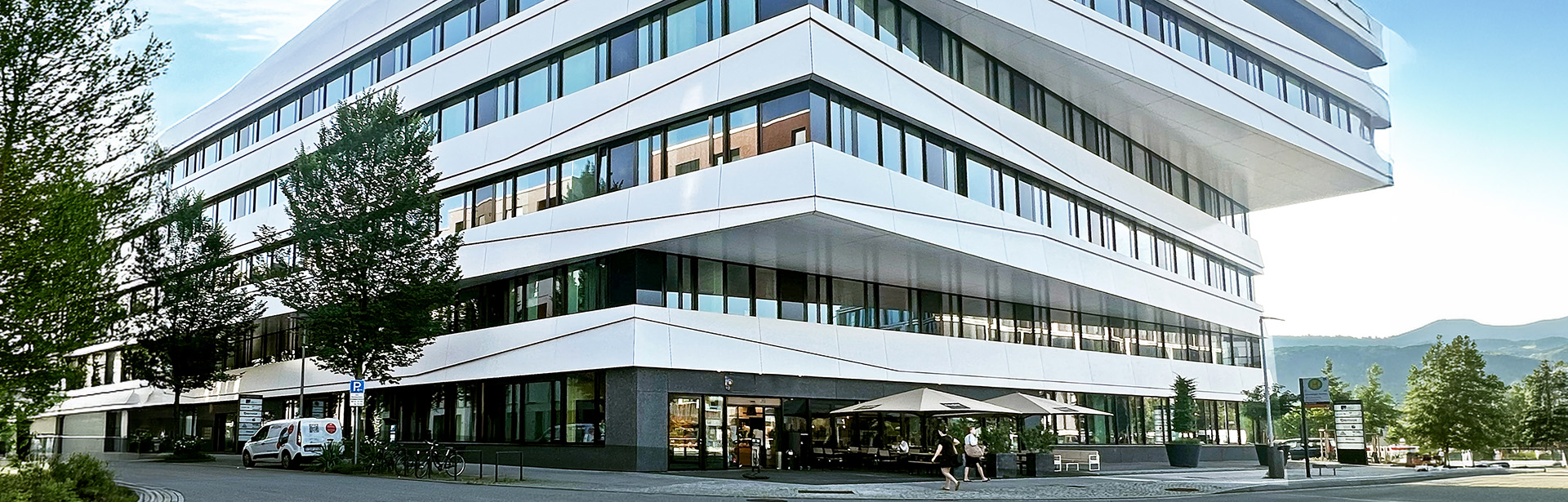 A modern building of io with a sleek, angular design features large windows and a shaded outdoor seating area.