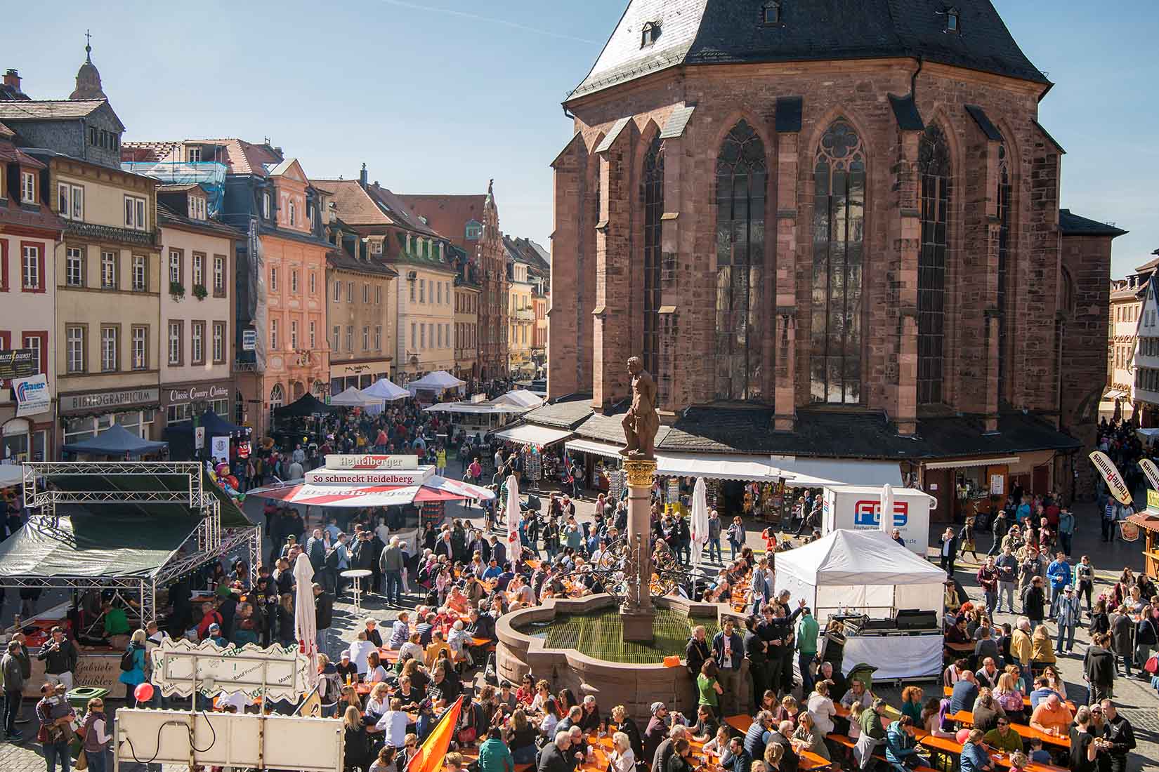 The bustling marketplace of Heidelberg filled with people enjoying food and drinks, surrounded by historic buildings and a large church.