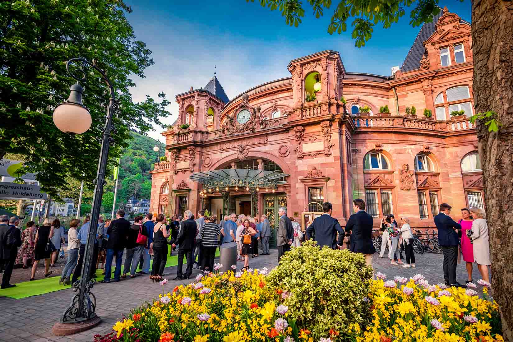The entrance of a historic municipal hall of Heidelberg buzzes with activity as attendees socialize, framed by a backdrop of colorful floral displays.
