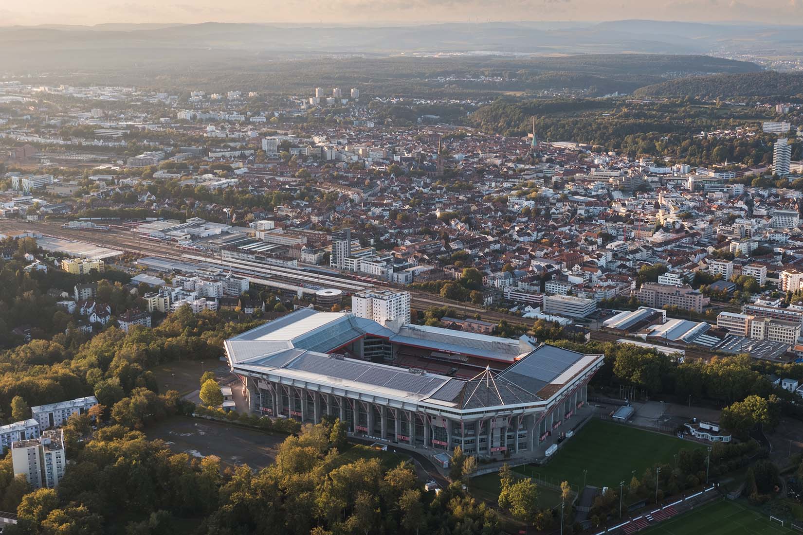 An aerial view captures a large stadium surrounded by lush trees and urban buildings, showcasing a vibrant cityscape.