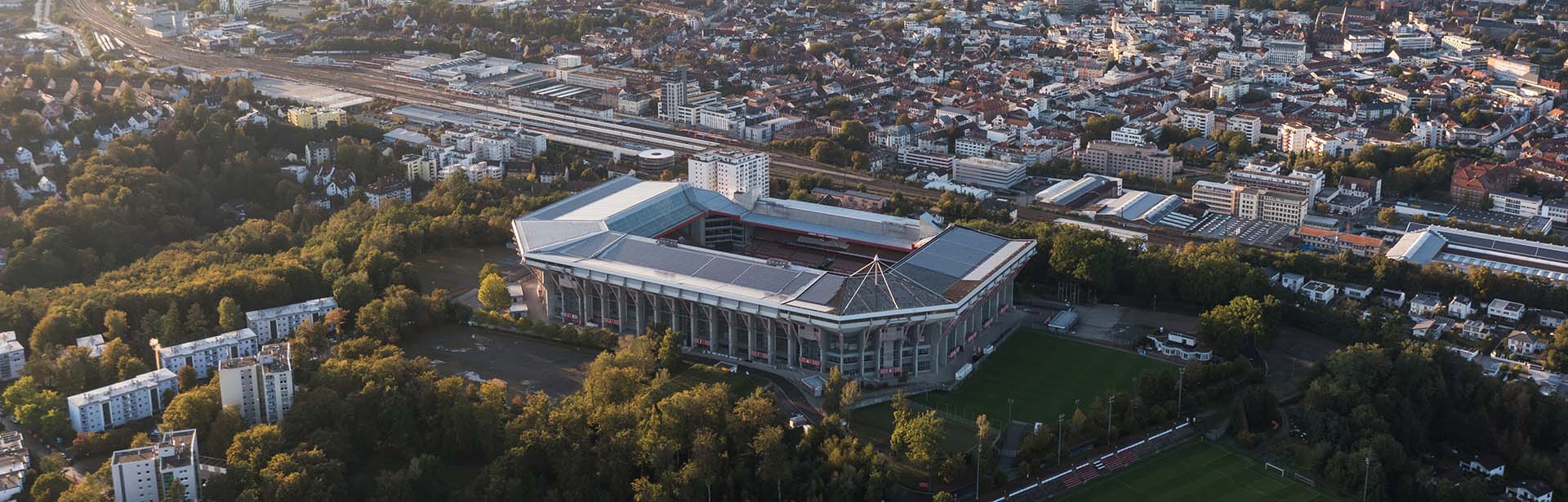 An aerial view captures a large stadium surrounded by lush trees and urban buildings, showcasing a vibrant cityscape.