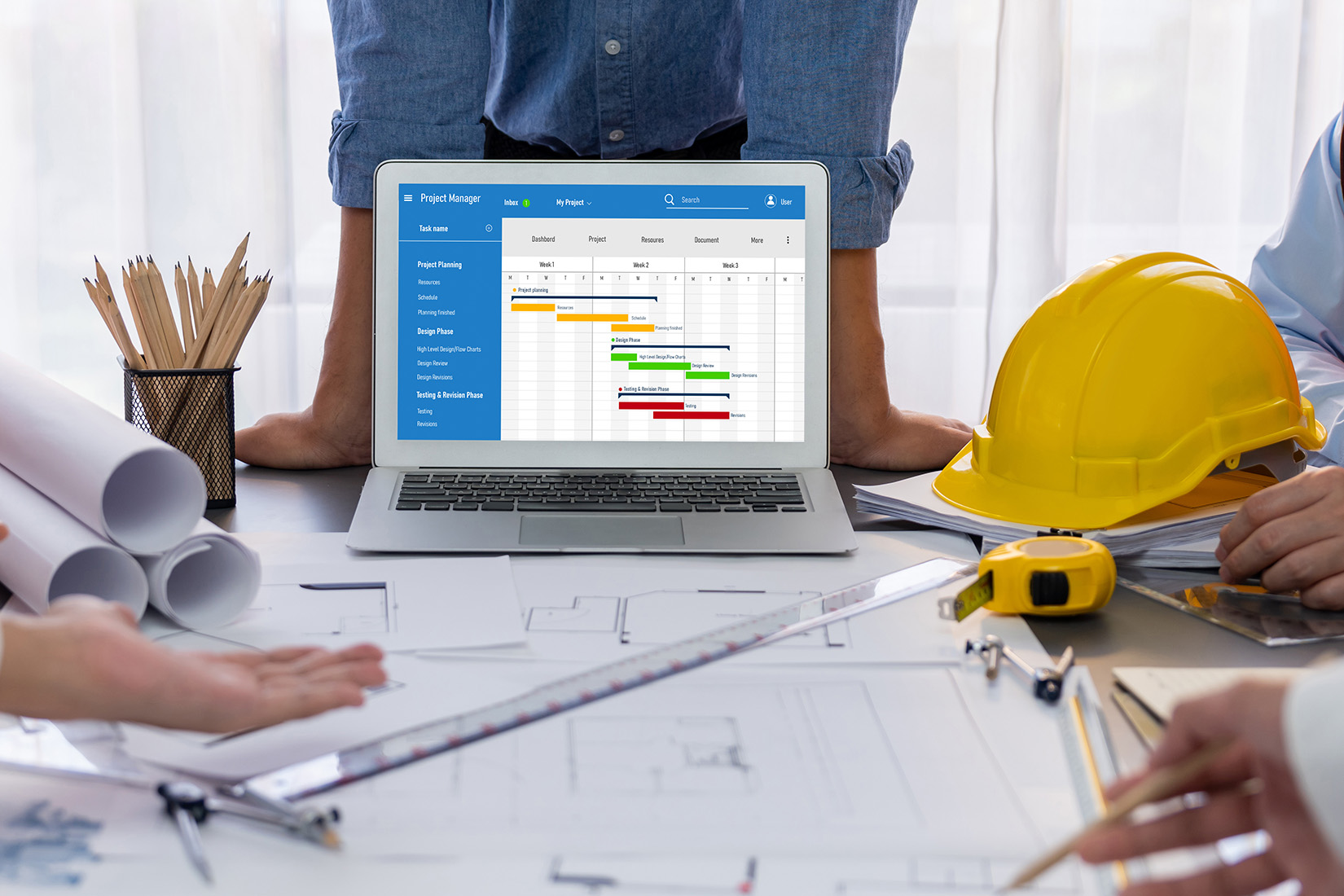 Blick auf ein Team, das intensiv an einem Projekt arbeitet, mit Bauplänen und einem Laptop, der einen detaillierten Zeitplan zeigt.