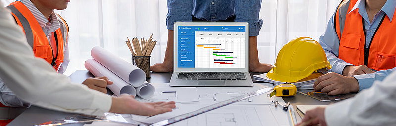 Blick auf ein Team, das intensiv an einem Projekt arbeitet, mit Bauplänen und einem Laptop, der einen detaillierten Zeitplan zeigt.